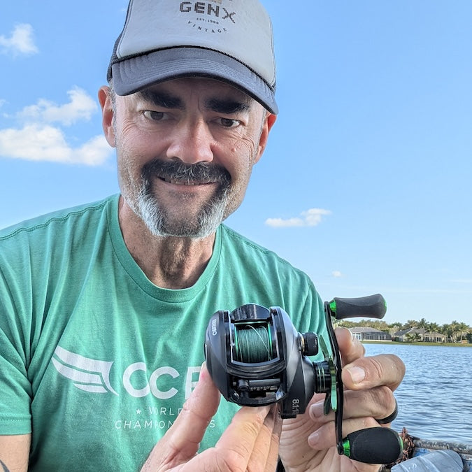 Man holding a fishing reel by a lake, wearing a green shirt and gray cap.