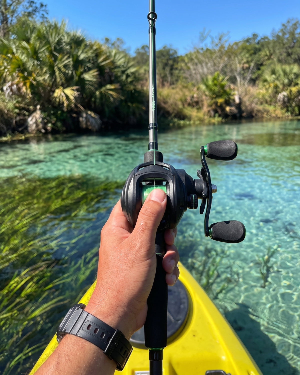 Close-up of a hand holding a casting rod with an Obalus baitcast reel over water, thumb on the spool