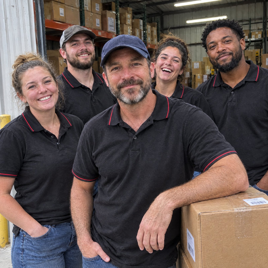 The Obalus team in black polo shirts at the open door of their warehouse, smiling at the camera, the owner leaning on a stack of shipping boxes