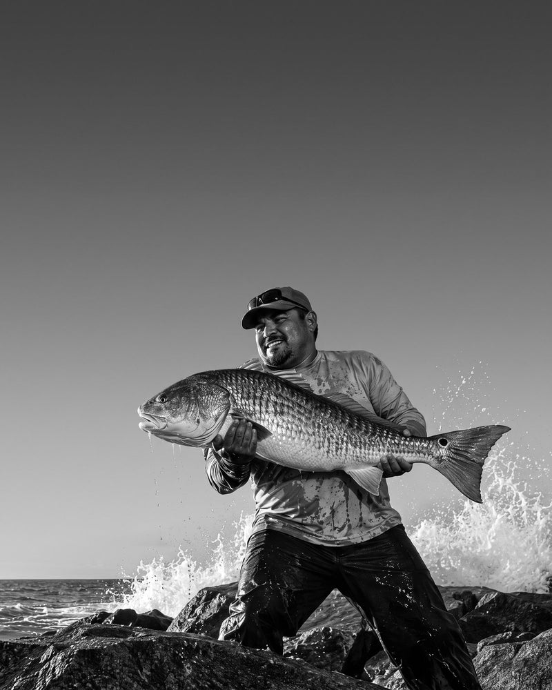 Black-and-white photo of an angler on Gulf coast jetty rocks lifting a big redfish as a wave breaks behind him