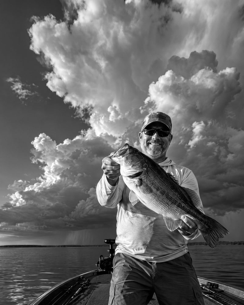 Black-and-white photo of a father kneeling on a pier at sunrise, helping his young son fight a fish